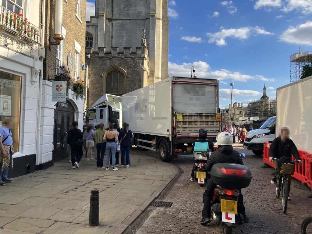 Photo of a street corner in a historic city centre with narrow streets. An articulated lorry is navigating the corner, using a little bit of the footway to help, with pedestrians watching the driver's efforts or perhaps just waiting to cross the street. Another lorry is waiting nearby. Several powered and unpowered two-wheelers, one of whom is fairly obviously a food delivery rider, are also in the mix.
