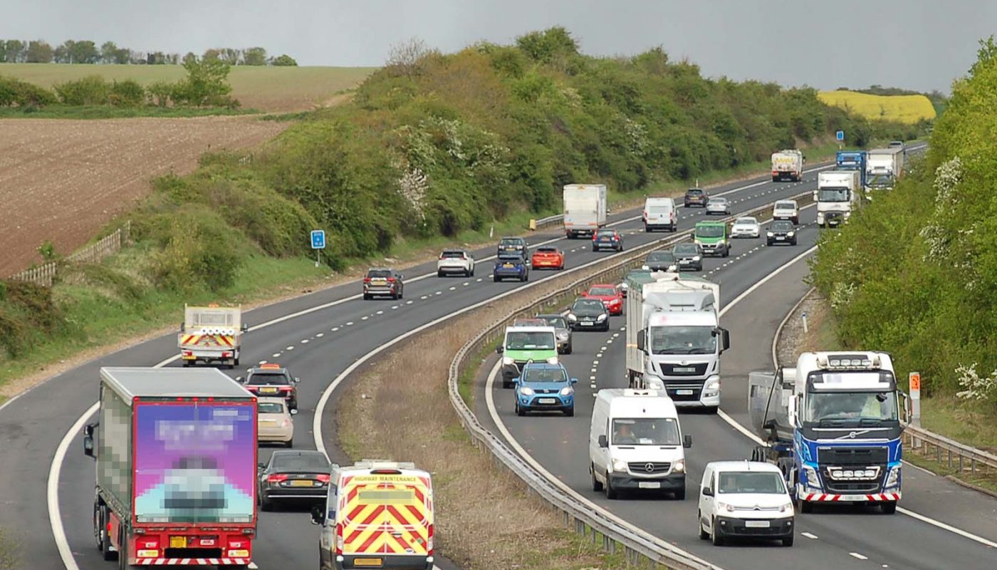 Photo looking along a dual two-lane motorway going around a curve and into a cutting in a rural arable area, under a grey sky. The road is quite busy with colourful traffic including quite a high proportion of vans and lorries.