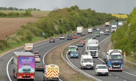 Photo looking along a dual two-lane motorway going around a curve and into a cutting in a rural arable area, under a grey sky. The road is quite busy with colourful traffic including quite a high proportion of vans and lorries.