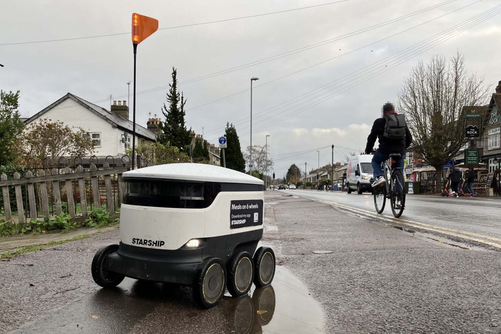 A 'starship' delivery robot on a wet footway on a wet day, on a local main road with a mix of commercial and residential frontages. A cyclist and a small freight van are passing by.