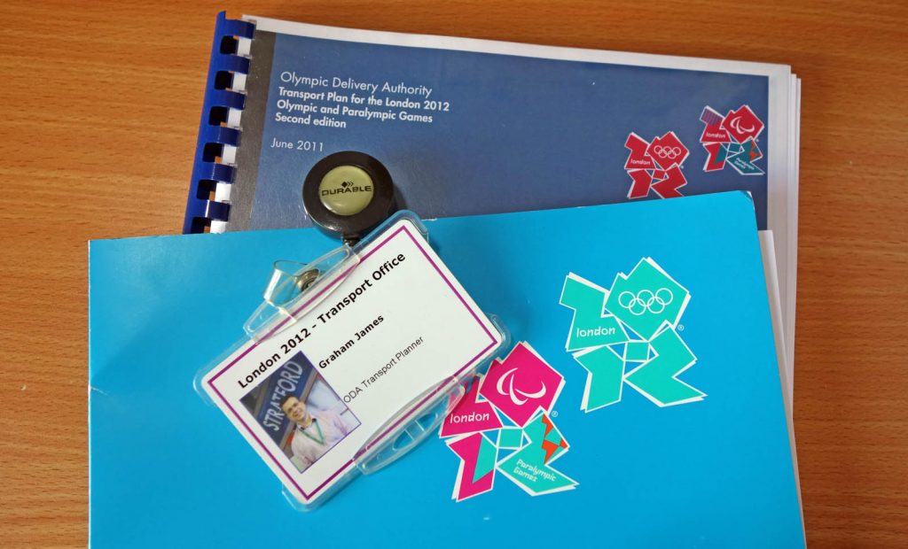 An ID badge, a cardboard folder, and a copy of the London 2012 transport plan, resting on a wood-effect desktop. The ID badge says “London 2012 – transport office. Graham James. ODA Transport Planner.” It includes a picture of Graham, smiling happily, in front of an Underground roundel saying ‘Stratford’. It is attached to some sort of retractable chain thing that clips onto a belt. The cardboard folder is turquoise-cyan with the London 2012 Olympic and Paralympic Games logos on it. The transport plan is comb-bound and looks like an office printout. We can see its front cover which also has the two logos, plus text reading “Olympic Delivery Authority. Transport Plan for the London 2012 Olympic and Paralympic Games. Second edition. June 2011.”