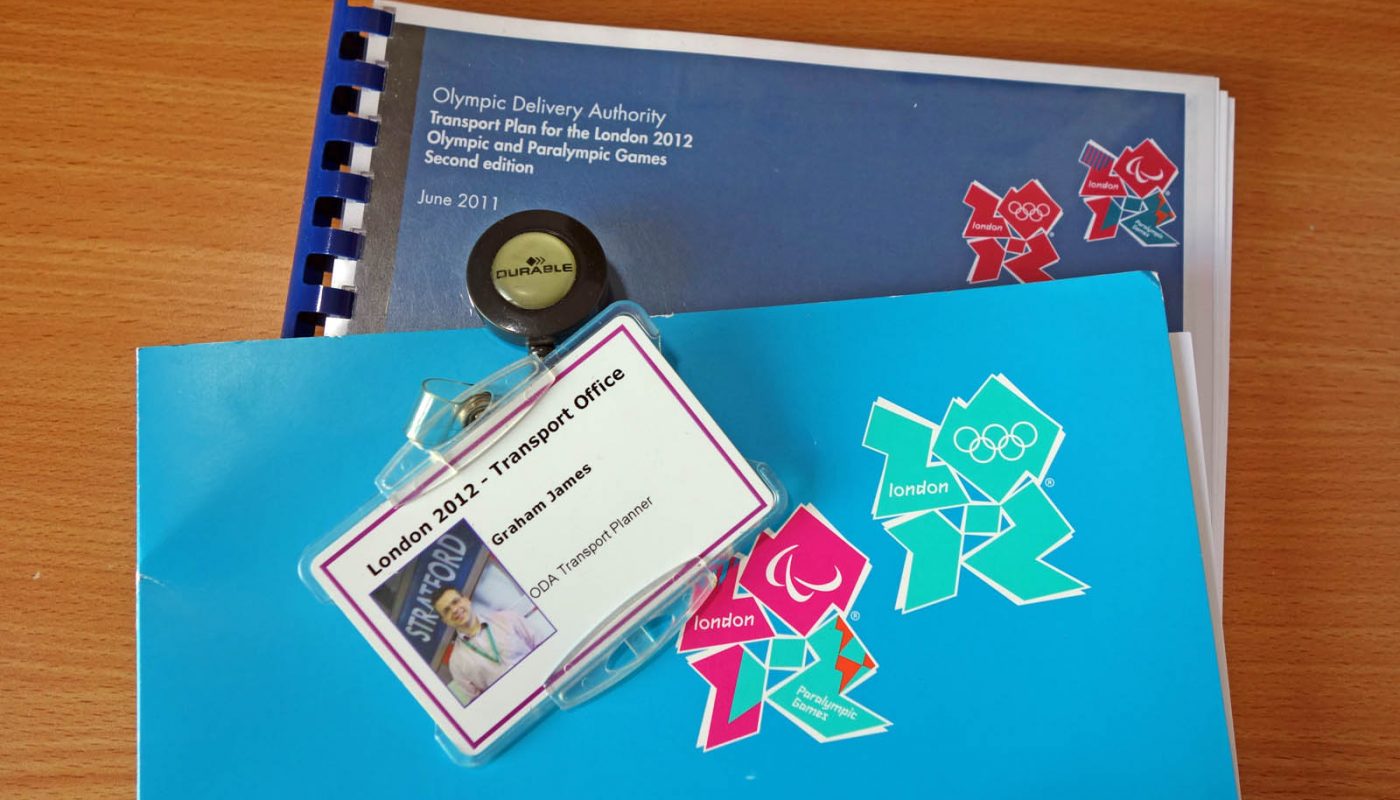 An ID badge, a cardboard folder, and a copy of the London 2012 transport plan, resting on a wood-effect desktop. The ID badge says “London 2012 – transport office. Graham James. ODA Transport Planner.” It includes a picture of Graham, smiling happily, in front of an Underground roundel saying ‘Stratford’. It is attached to some sort of retractable chain thing that clips onto a belt. The cardboard folder is turquoise-cyan with the London 2012 Olympic and Paralympic Games logos on it. The transport plan is comb-bound and looks like an office printout. We can see its front cover which also has the two logos, plus text reading “Olympic Delivery Authority. Transport Plan for the London 2012 Olympic and Paralympic Games. Second edition. June 2011.”