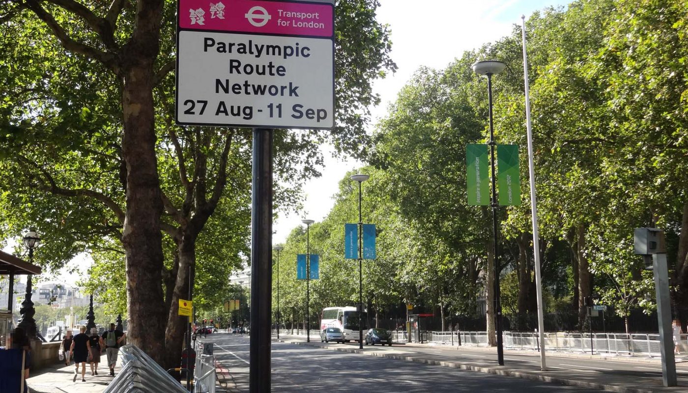 Photo looking along the Embankment (a major road in central London). A sign, with the London 2012 logo, says that it is part of the Paralympic Route Network. There are crowd barriers along the edge of the footway. People are walking. Traffic is light.