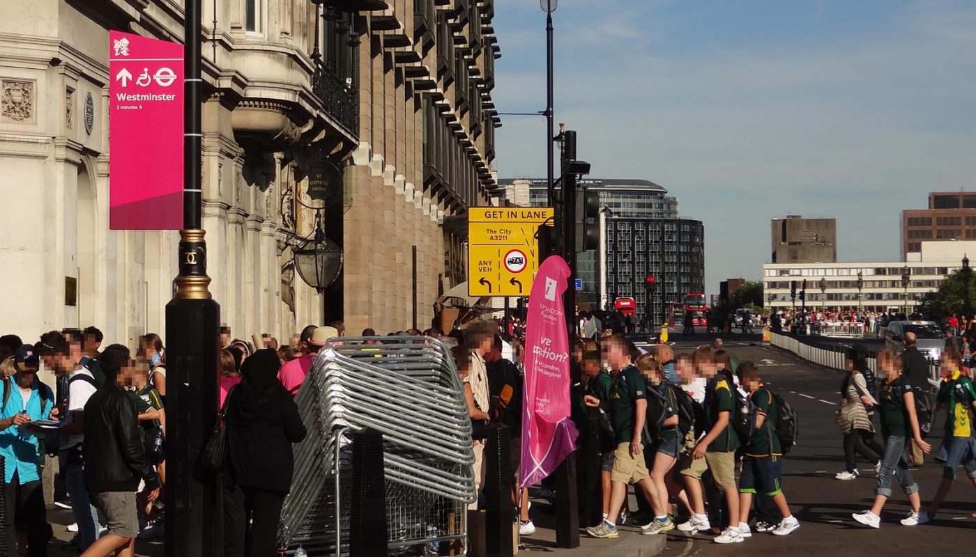 Photo of a street in central London during the London 2012 Games. The street is busy with people. A magenta banner on a lamp column is giving directions to Westminster station, two minutes' walk away. We can just see a Games Ambassador, behind a stack of crowd barriers that are not currently in use. In the background is a street with some buses and taxis but not much other motorised traffic.