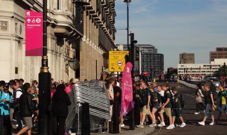 Photo of a street in central London during the London 2012 Games. The street is busy with people. A magenta banner on a lamp column is giving directions to Westminster station, two minutes' walk away. We can just see a Games Ambassador, behind a stack of crowd barriers that are not currently in use. In the background is a street with some buses and taxis but not much other motorised traffic.