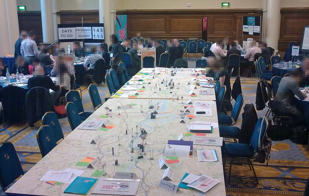 Photo showing a large room with lots of tables and chairs, and people in business clothes. In the centre, dominating the room, is a very large table. It has about twenty chairs around it, and a map of central London as the tabletop. There are 'game pieces' dotted around in the shape of major landmarks and with the symbols of different transport networks. Various papers are on the edge of the table in front of the chairs. We can see that the one nearest to us is a bound A4 set of papers leaded 'Transport Exercise 3: Scenario 1 and 2 Map Pack". We can also see cardboard folders in aquamarine colour with the London 2012 logos on them, and sets of A6-size red, orange and green cards. In the corner of the room, a large notice-board tells us that there are 148 days to go until the Paralympics, and 115 until the Olympics.