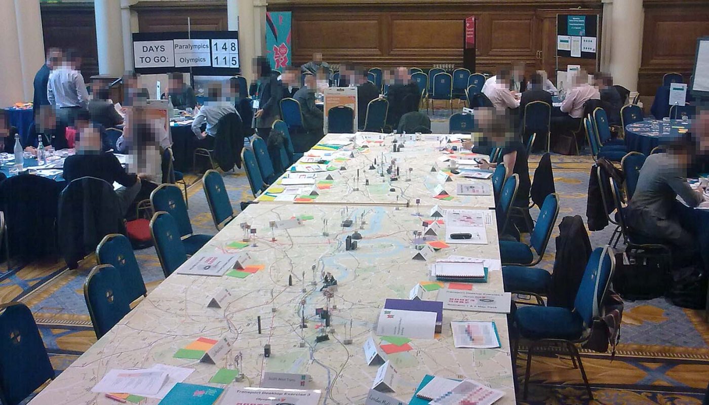 Photo showing a large room with lots of tables and chairs, and people in business clothes. In the centre, dominating the room, is a very large table. It has about twenty chairs around it, and a map of central London as the tabletop. There are 'game pieces' dotted around in the shape of major landmarks and with the symbols of different transport networks. Various papers are on the edge of the table in front of the chairs. We can see that the one nearest to us is a bound A4 set of papers leaded 'Transport Exercise 3: Scenario 1 and 2 Map Pack". We can also see cardboard folders in aquamarine colour with the London 2012 logos on them, and sets of A6-size red, orange and green cards. In the corner of the room, a large notice-board tells us that there are 148 days to go until the Paralympics, and 115 until the Olympics.
