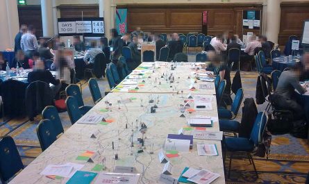 Photo showing a large room with lots of tables and chairs, and people in business clothes. In the centre, dominating the room, is a very large table. It has about twenty chairs around it, and a map of central London as the tabletop. There are 'game pieces' dotted around in the shape of major landmarks and with the symbols of different transport networks. Various papers are on the edge of the table in front of the chairs. We can see that the one nearest to us is a bound A4 set of papers leaded 'Transport Exercise 3: Scenario 1 and 2 Map Pack". We can also see cardboard folders in aquamarine colour with the London 2012 logos on them, and sets of A6-size red, orange and green cards. In the corner of the room, a large notice-board tells us that there are 148 days to go until the Paralympics, and 115 until the Olympics.