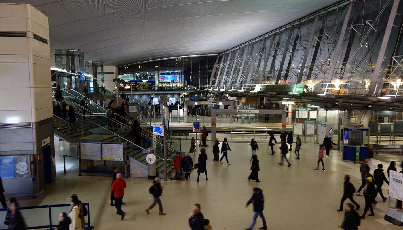 Photo inside the main ticket hall of Stratford station, after dark, looking from the first floor at one end, over the Jubilee line concourse and across the DLR platforms towards the main gateline at the other end. The ticket hall is busy with people.