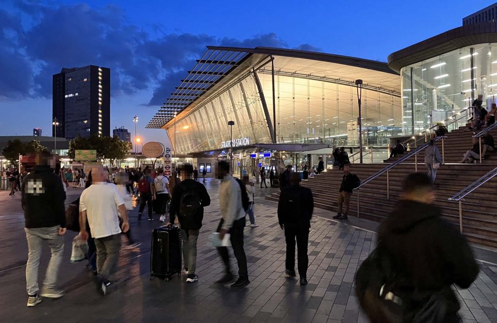 Photo on the plaza outside Stratford station at dusk, looking towards the main ticket hall and the foot of the steps towards Westfield. The area is quite busy with people. The sky is a dramatic blue,, with the inside of the ticket hall contrasting in a yellow light seen through the high glass walls.