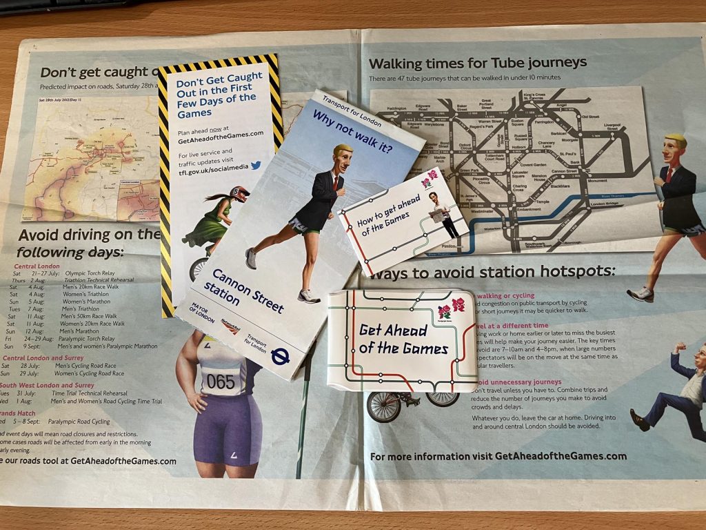A collection of London 2012 'Get ahead of the Games' travel demand management (TDM) items. It includes a double-page newspaper advert, a leaflet headlined 'Don't get caught out in the first few days of the Games', a folded walking map leaded "Why not walk it?" for Cannon Street station, a credit-card sized foldout leaflet titled 'How to get ahead of the Games', and an Oyster card wallet headed 'Get Ahead of the Games'. The items feature cartoon-style illustrations, such as a man wearing a shirt, tie and jacket with shorts and trainers who is striding purposefully.