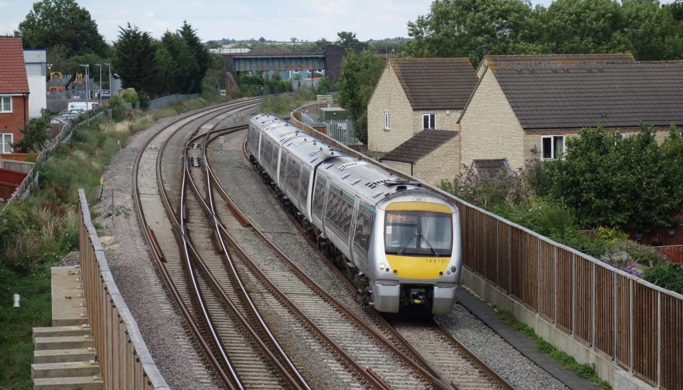 Photo of a railway line at a double junction, in the facing direction as seen from the camera. There are modern houses on either side of the line, with noise barriers shielding them from the railway. A predominantly silver-coloured diesel multiple unit is crossing the junction, coming towards the camera on the right-hand-most track.
