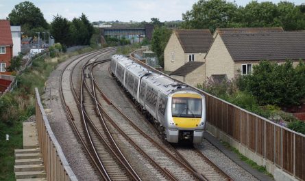 Photo of a railway line at a double junction, in the facing direction as seen from the camera. There are modern houses on either side of the line, with noise barriers shielding them from the railway. A predominantly silver-coloured diesel multiple unit is crossing the junction, coming towards the camera on the right-hand-most track.
