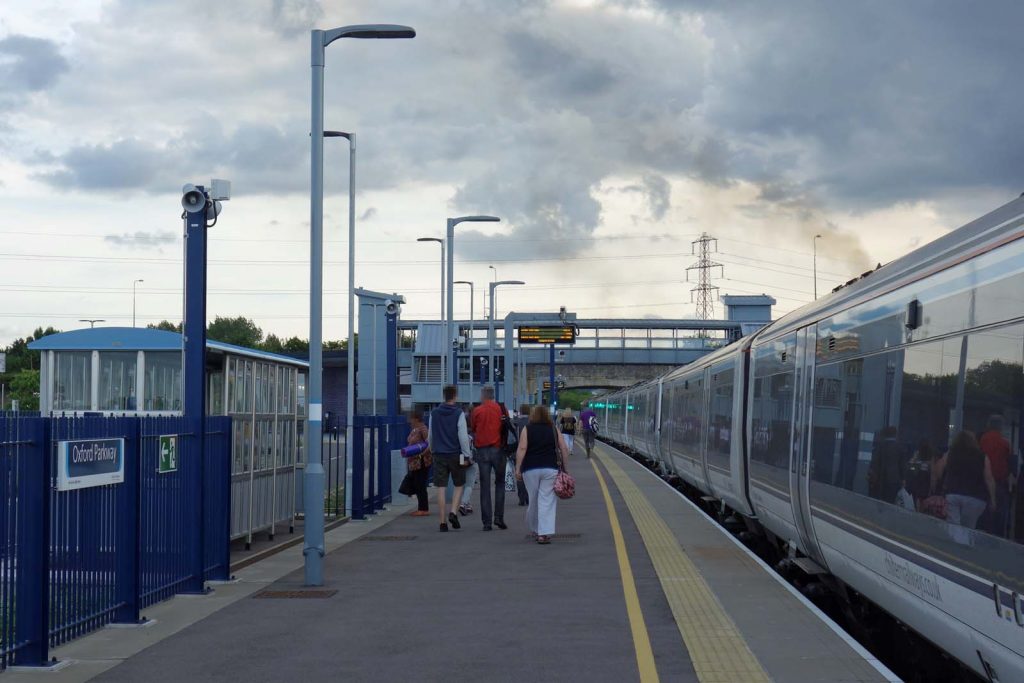 Photo looking along a station platform. It looks like quite a modern station., There is a train at the platform and people are walking along the platform away from the camera.
