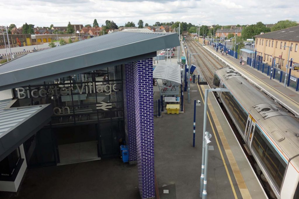 Photo looking along a railway station platform from an elevated position. It is a two-track railway with side platforms at this urban station. A diesel multiple unit is at the platform below the camera. This platform has a striking modern station building, with a two-storey concourse, sloping roofs and distinctive external columns of what look like blue glazed bricks with white mortar. The station name 'Bicester Village' is seen on the side of the building.