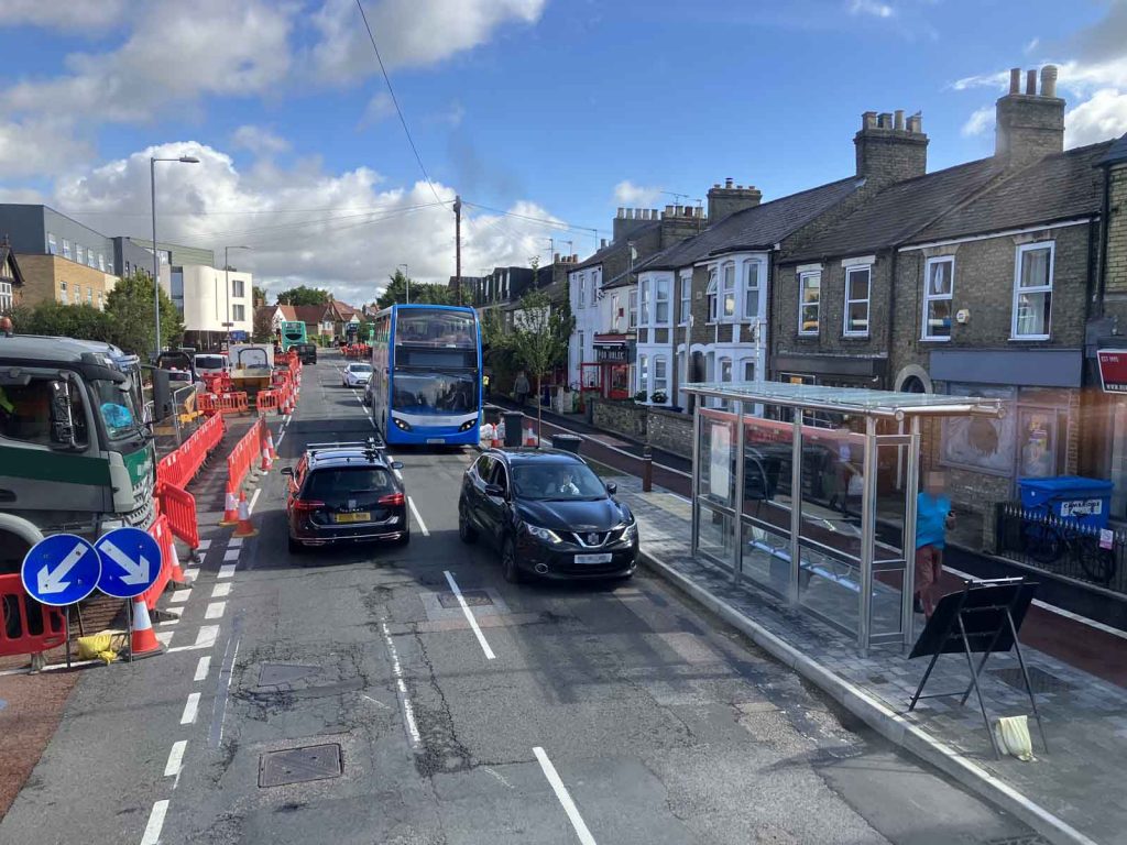 An urban arterial street, with construction work in progress. There is what looks like a new cycle track and a new bus stop boarder with shelter. Traffic is going past, including a bus. There is a lorry on the left, within the barriered-off construction area.