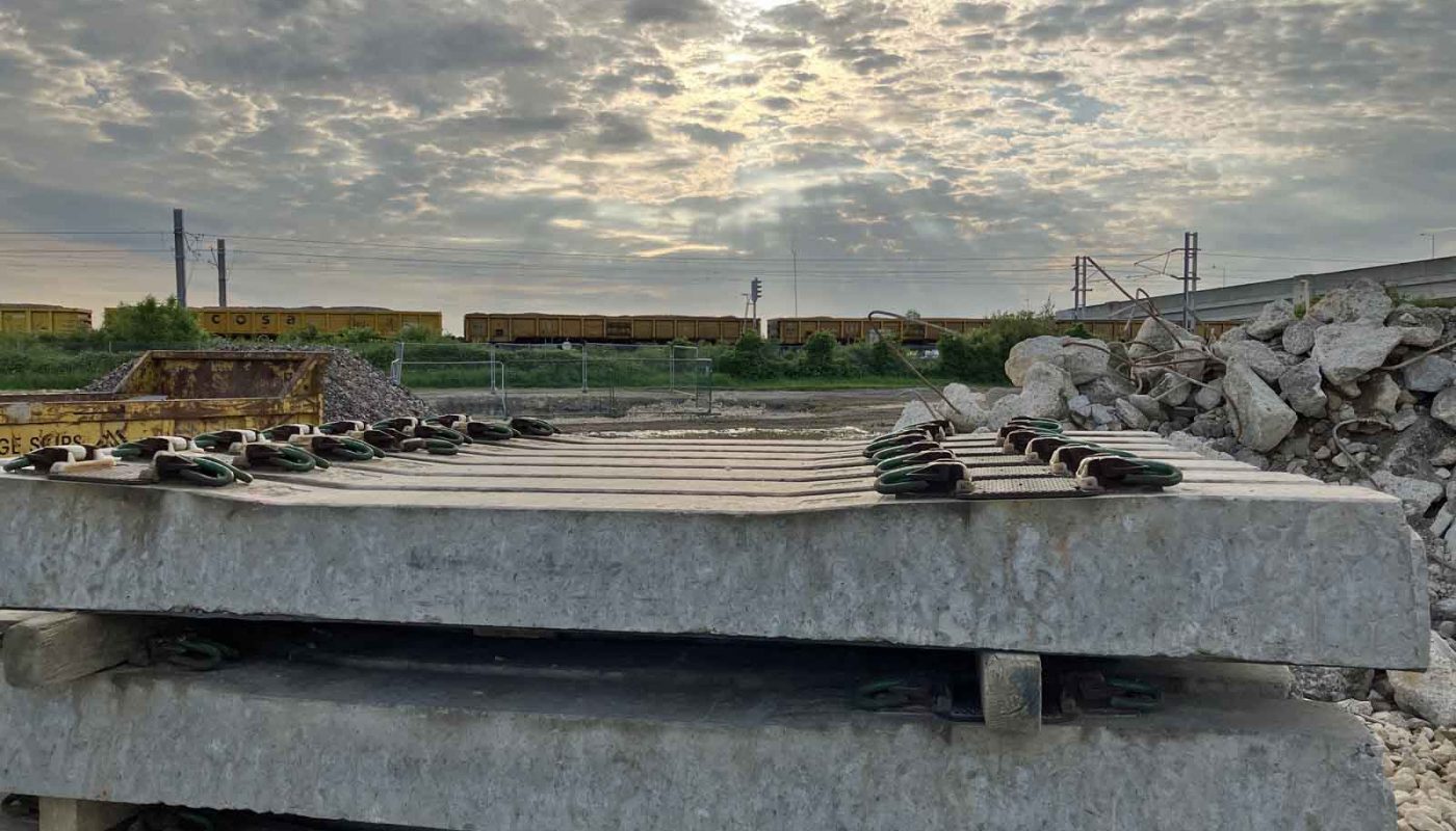 Photo looking at a construction compound with a stack of concrete sleepers in the foreground. There is also a pile of reinforced-concrete rubble. In the background is a double-track overhead-electrified railway line, with a rake of yellow box wagons filled with aggregates.