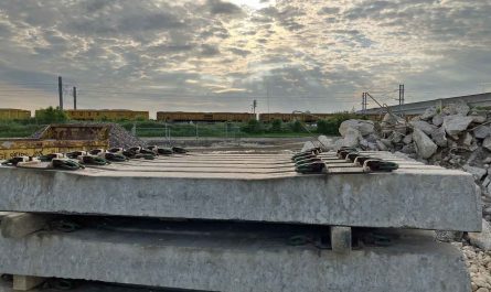 Photo looking at a construction compound with a stack of concrete sleepers in the foreground. There is also a pile of reinforced-concrete rubble. In the background is a double-track overhead-electrified railway line, with a rake of yellow box wagons filled with aggregates.