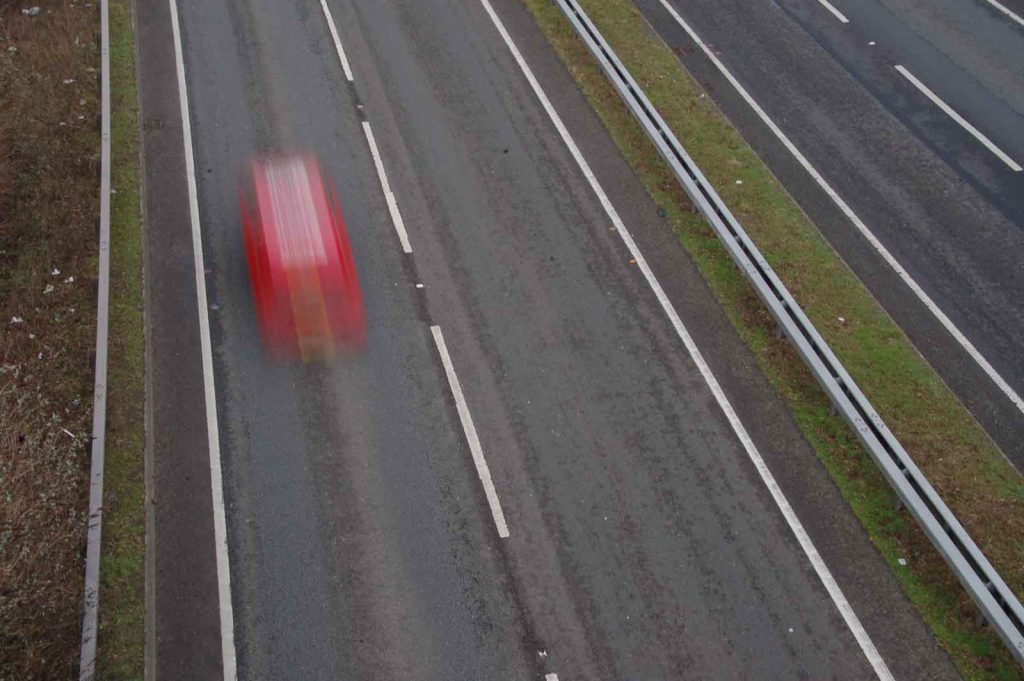 Photo looking down on a dual-carriageway road, with a blurred red vehicle, probably a car, going past at speed.