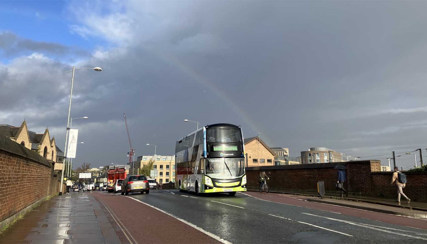 Photo of a street, with various people using various modes of transport, under a mostly grey, partly blue sky, with a rainbow.