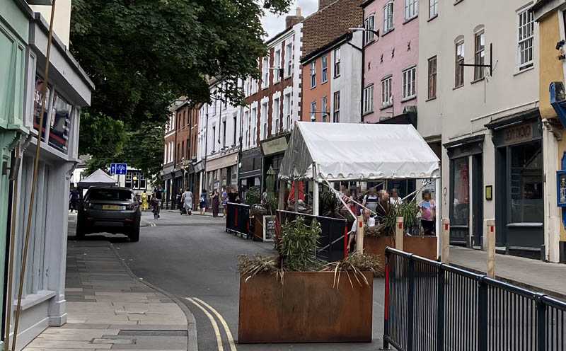 A city street, with traditional buildings with shopfronts. Some of the carriageway space has been given over to dining areas. It looks rather pleasant.