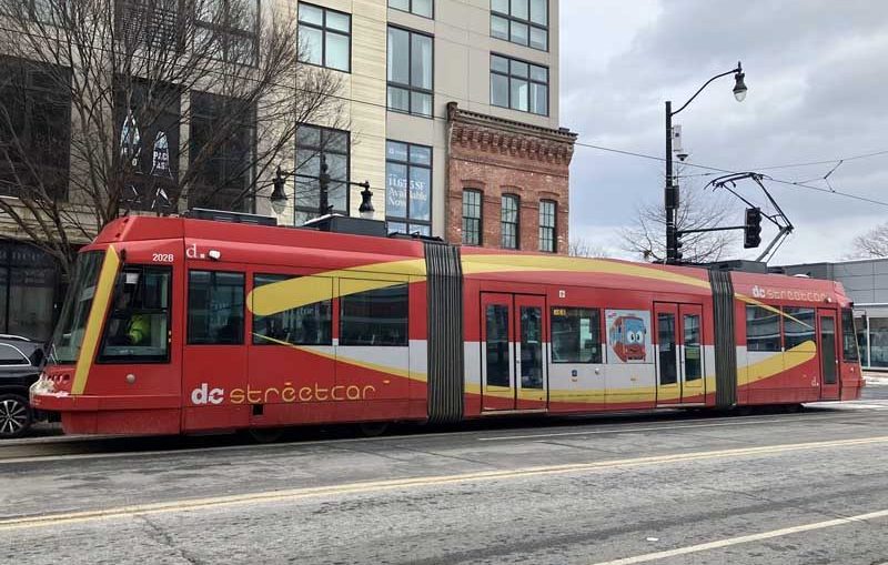 Photo looking across a city street to a streetcar vehicle. The vehicle is double-articulated, in predominantly red livery, and says "DC Streetcar"