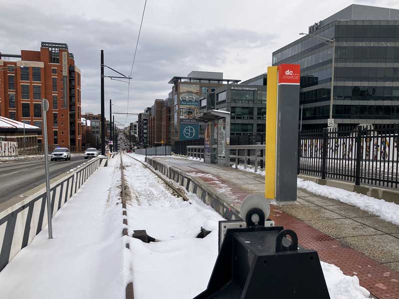 Photo looking down the track from the bumper (buffer-stop) at the start/finish of an on-street urban streetcar line. We are in the middle of a road. There is a single track, with trolley wiring, and a raised platform alongside for the stop. There is snow on the ground, which has been cleared from the platform and the road carriageways.