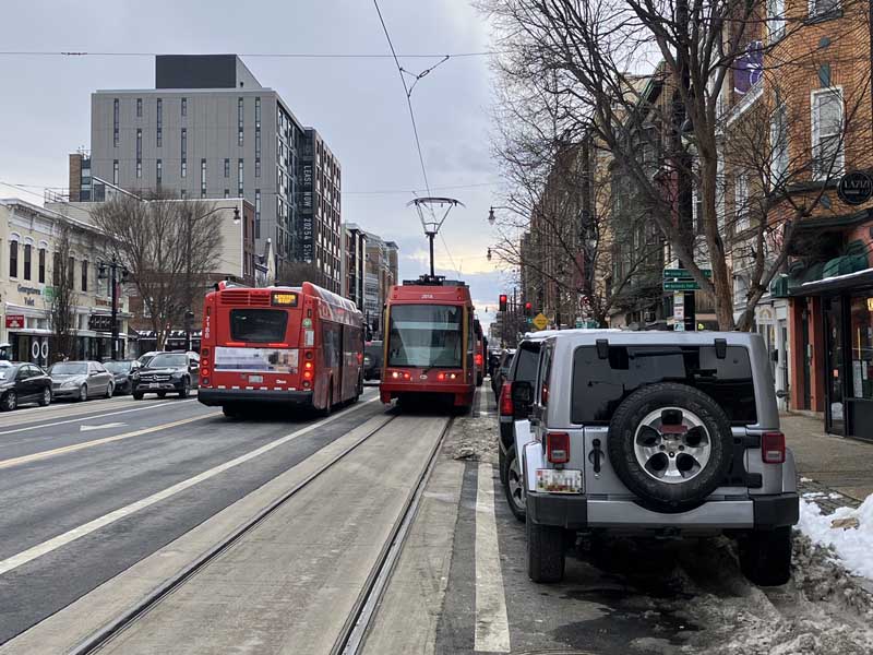 VIew along a city street with shopfronts. Cars are parked alongside the kerb. Next to the cars, there is a streetcar track with a concrete trackbase and trolley wiring. A red streetcar vehicle is in the middle of the picture. In the adjacent traffic lane, a red single-decker bus is passing the streetcar vehicle. There is some snow or slush on the ground, mostly  in the gutter areas of the parking and the adjoining part of the sidewalk (footway).