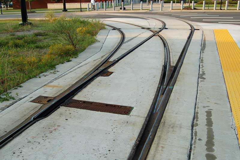 Photo of a switch (point) in tramway track, where two tracks become one. We are at the toe end (one track), looking towards the heel (two tracks). The switch is in a concrete track base, alongside a street. The track at the toe end is perpendicular to the street. The diverging route is to the left. Both routes immediately take a sharp right-angle turn left onto the street. The switch has only one switch rail, currently set for the straight-ahead route. On the other toe, where the switch rail would have been, the groove is raised and has bright contact paths on both routes through it, indicating flange-running. Elsewhere on the track, there are bright contact paths on various different areas of the railheads.