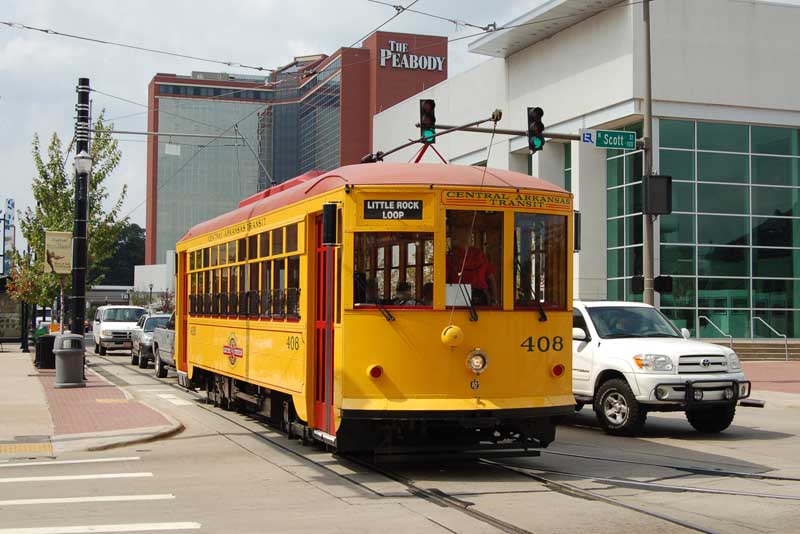 Photo at a signalised crossroads of city streets. Coming towards the camera is an old-fashioned-looking (but modern) streetcar. It has just taken the straight-ahead route over a switch (points) which offers a diverging route to the left (right of photo). There is other traffic around the streetcar, including SUV-style pickup trucks. The streetcar is in predominantly yellow livery with red doors and roof. It has the name and logotype of Central Arkansas Transit and is numbered 408. The destination blind says "Little Rock Loop". The streetcar is collecting current from the overhead trolley wire using its rear trolley connector; we can also see the lowered front trolley connector.