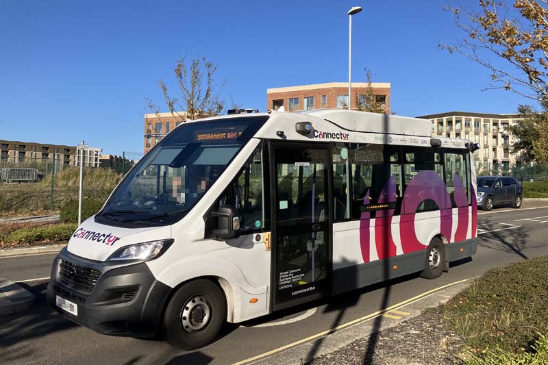 Photo of a midibus en route on a road on a sunny day with long shadows. The road looks like it’s in a large recent development. The bus is in a predominantly white livery, branded as ‘Connector’. Under the usual legal lettering on the side, it says ‘Autonomous Bus’. There’s a person in the driving seat (pixelated in this photo).