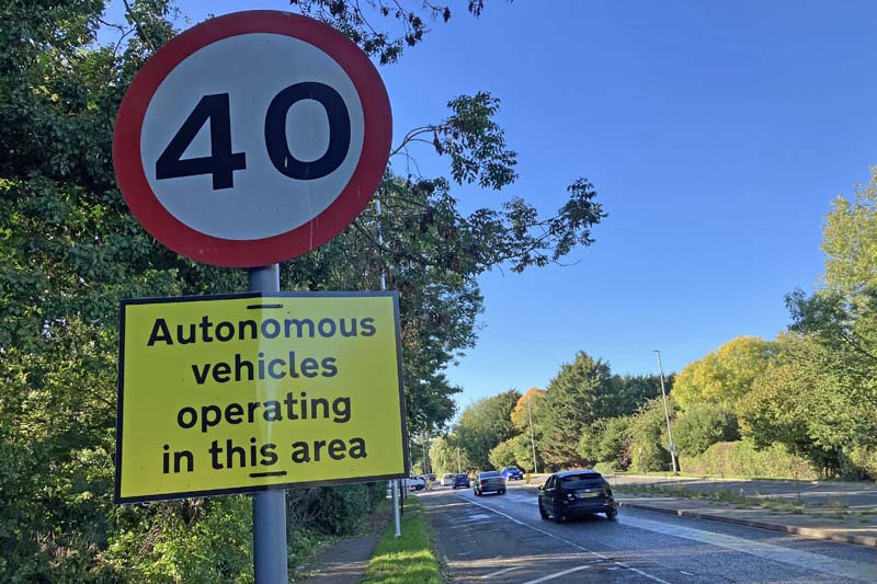 Photo looking along an arterial road on a sunny day. In the foreground is a 40mph speed limit sign. Attached to the pole is a temporary sign, with black text on a yellow background, reading "Autonomous vehicles operating in this area". The road has trees and hedges on each side. Cars are being driven along the road.