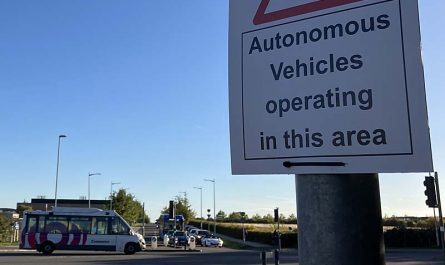 Photo looking across a signalised road junction on a sunny day. There are few buildings visible and the junction looks rural or at least on the edge of an urban area. In the foreground, a sign reading ‘Autonomous vehicles operating in this area’ is attached to a pole. In the background, a midibus is crossing the junction from left to right. It is in predominantly white livery and says ‘Connector’ on it. Beyond, a queue of traffic on the side road includes some cars and a single-decker bus.