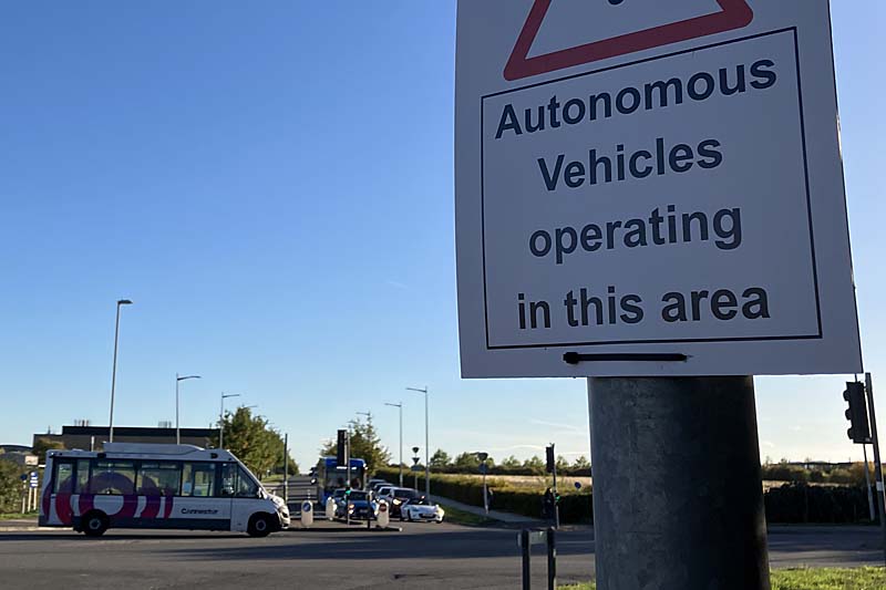 Photo looking across a signalised road junction on a sunny day. There are few buildings visible and the junction looks rural or at least on the edge of an urban area. In the foreground, a sign reading ‘Autonomous vehicles operating in this area’ is attached to a pole. In the background, a midibus is crossing the junction from left to right. It is in predominantly white livery and says ‘Connector’ on it. Beyond, a queue of traffic on the side road includes some cars and a single-decker bus.
