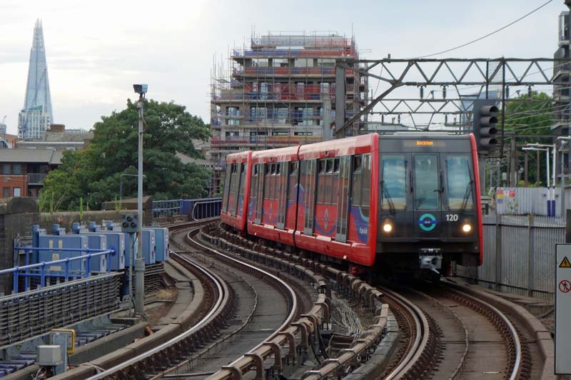 Photo looking along an urban railway line on a viaduct. A train is approaching on one of the two tracks. It is composed of two (or maybe more) urban metro type units, in predominantly red livery with "DLR" roundels. The destination display reads 'Beckton'. The line has bottom-contact third rail current collection. Alongside, there are elements of what seems to be another railway line; this one looks more main-line with its overhead elctrification gantries and four-aspect signalling. In the left background corner, a glass-faced office tower (triangular in profile) is in the distance, looking a bit like a shard of glass.