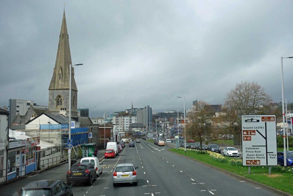 Photo on an overcast day, looking down an urban dual carriageway with shops on the left and a prominent church spire. A road sign shows directions to the City Museum, Continental ferry, Torpoint, Cattedown, Prince Rock, Queen Anne's Battery Marina, and Friary Park.