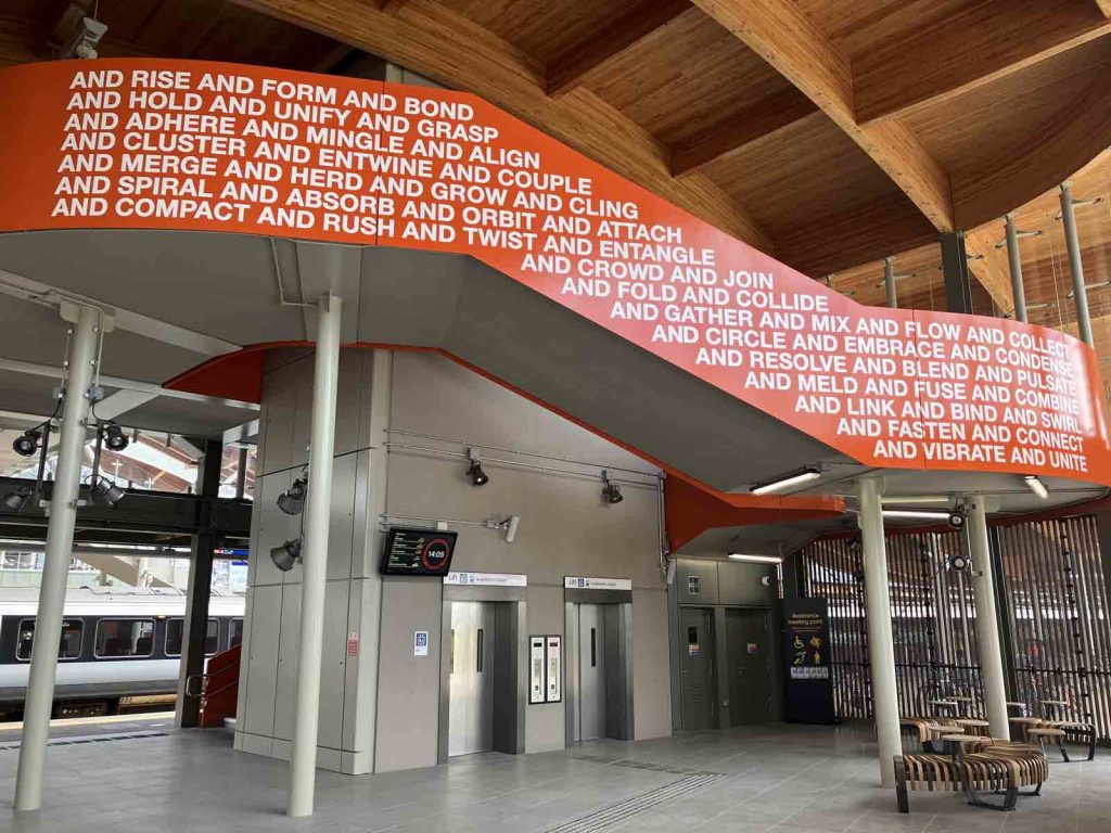 Photo inside the west side building at Cambridge South station, at ground level. We see the two lift doors, some benches, and the outer parapet of the staircase as it curls around the lift shafts. On the bright orange parapet, there is text: "And rise and form and bond and hold and unify and grasp and adhere and mingle and align and cluster and entwine and couple and merge and herd and grow and cling and spiral and absorb and orbit and attach and compat amd rush and twist and entangle and crowd and join and fold and collide and gather and mix and flow and collect and circle and embrace and condense and resolve and blend and pulsate and meld and fuse and combine and link and bind and swirl and fasten and connect and vibrate and unite".