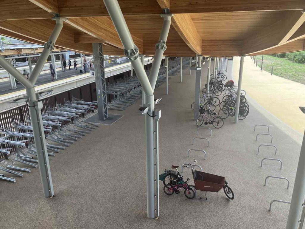 Photo looking down on the west side cycle parking area at Cambridge South. There are standard Sheffied stands, two-tier stands, and (nearest the camera) a set of widely-spaced, long-and-low Sheffield type stands. A Bakfiets cargo cycle and a child's cycle are attached to one of these stands. Other cycles, including one with a child trailer, are attached to the conventional Sheffield stands.