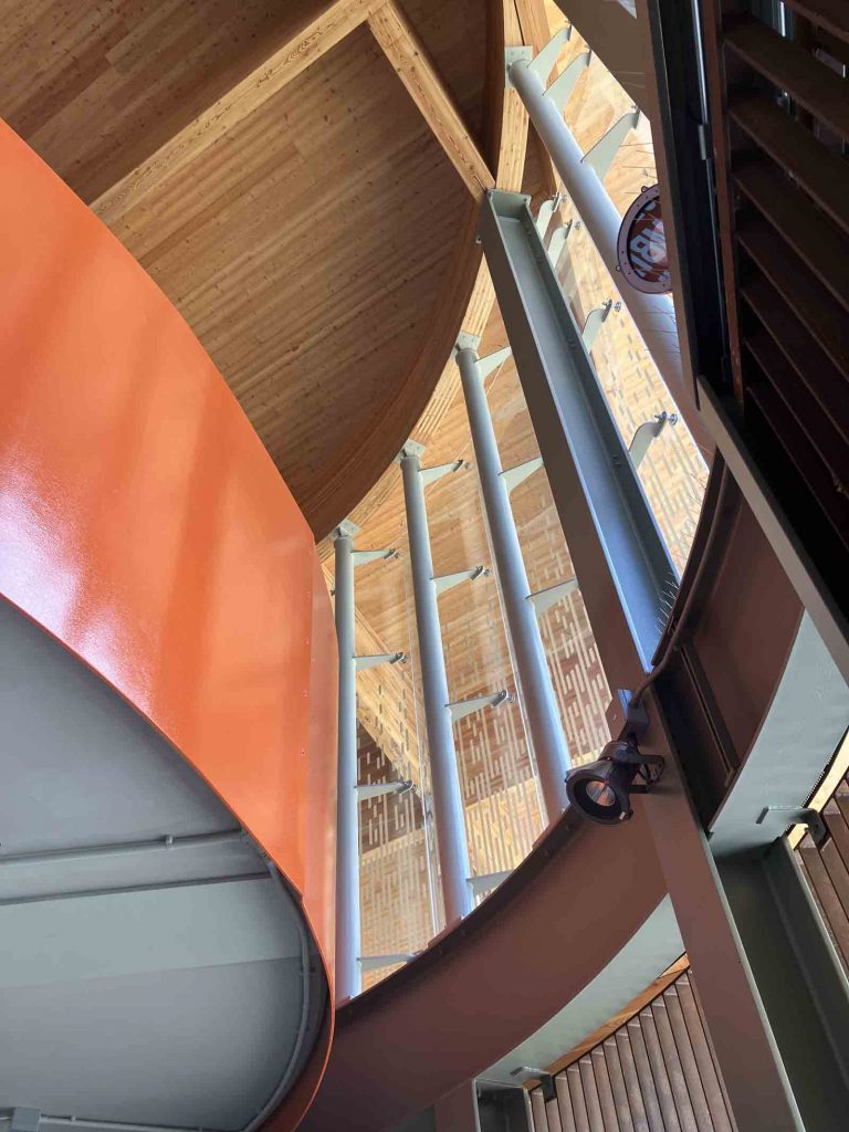 Photo looking upwards within one of the main buildings of Cambridge South, showing the orange staircase parapets and the engineered-timber interior of the roof.