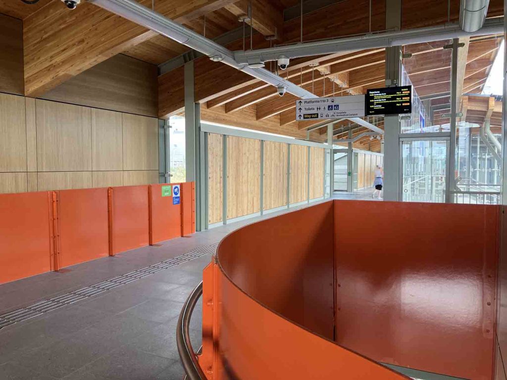 Photo taken from the top of one of the staircases on the main footbridge at Cambridge South station, looking towards the bridge. The parapets are steel panels, painted bright orange. There is a lot of exposed engineered timber too.