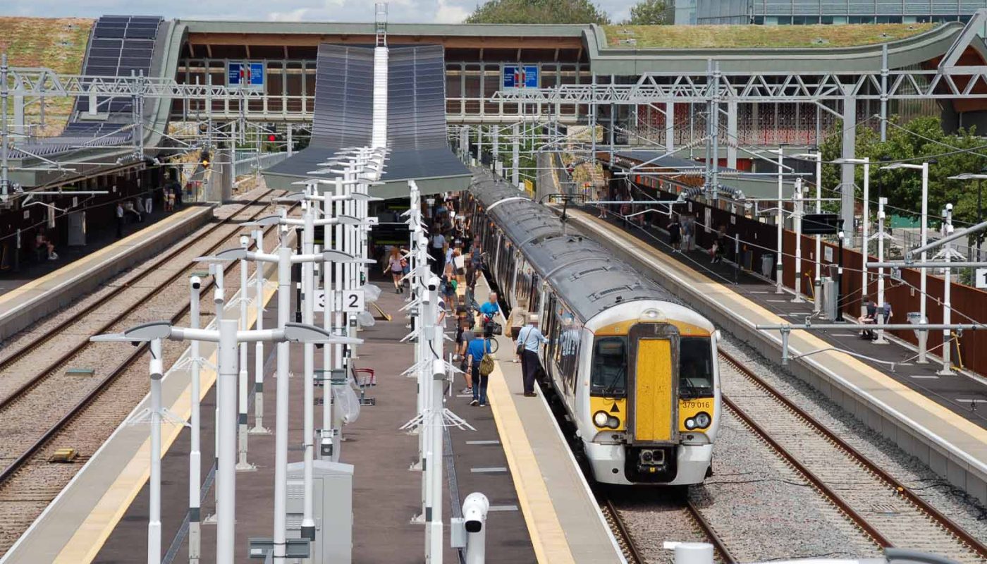 Photo taken from an elevated position, looking along the platforms at Cambridge North. A train of two class 379 units is stopped at platform 3. People are boarding and alighting. The main station building is in the background.
