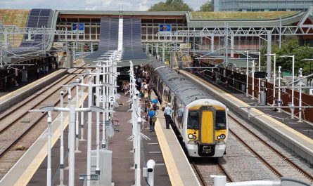 Photo taken from an elevated position, looking along the platforms at Cambridge North. A train of two class 379 units is stopped at platform 3. People are boarding and alighting. The main station building is in the background.