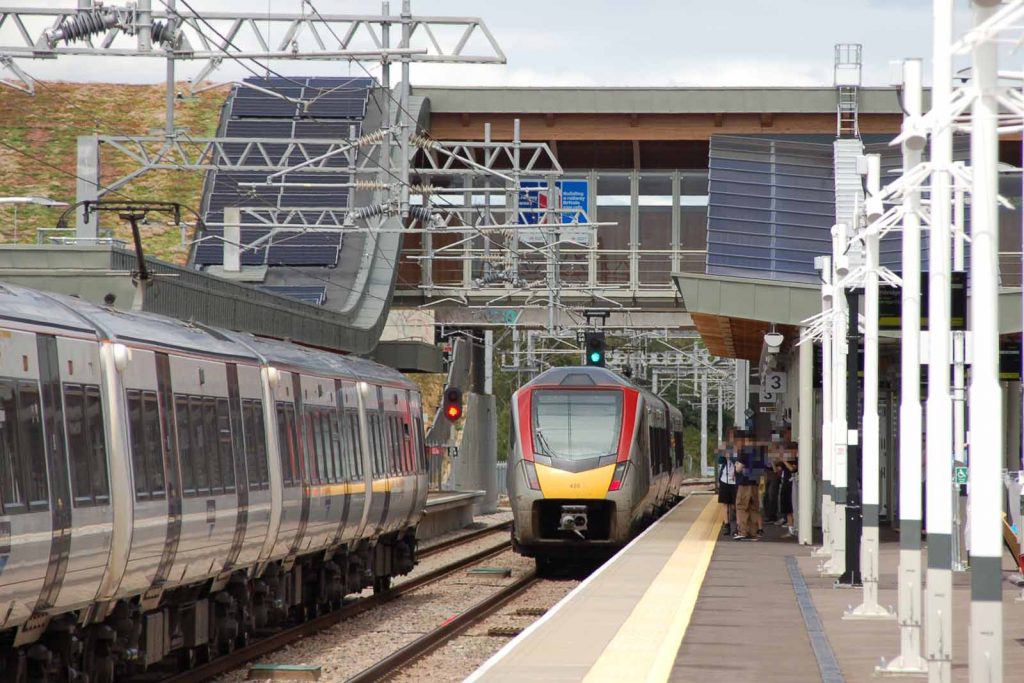 Photo looking north along platform 3 at Cambridge station, with platform 4 (the other down (northbound) platform on left of picture. A Greater Anglia Stadler unit has a green signal to leave platform 3. Next to it, a Great Northern electrostar unit is rolling into platform 3 on a red signal. Photo also shows a group of young enthusiasts enjoying the occasion with their cameras, the main footbridge and the grass roof of the west side main building.