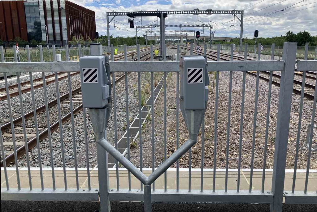 Photo at the platform end, looking towards the station starting signals. In the foreground, on the platform near the platform-end fence, are two signal post telephones sharing a common, Y-shaped galvanised steel pole.