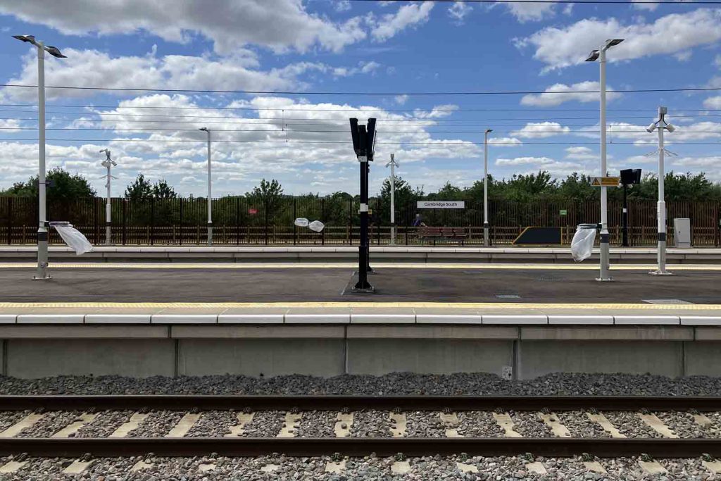 Photo looking across Cambridge South station from one of the side platforms, across the island platform, to the other side platform. The tall back-of-platform railings look unobtrusive in this picture, and we can see the trees and bushes in the park beyond.