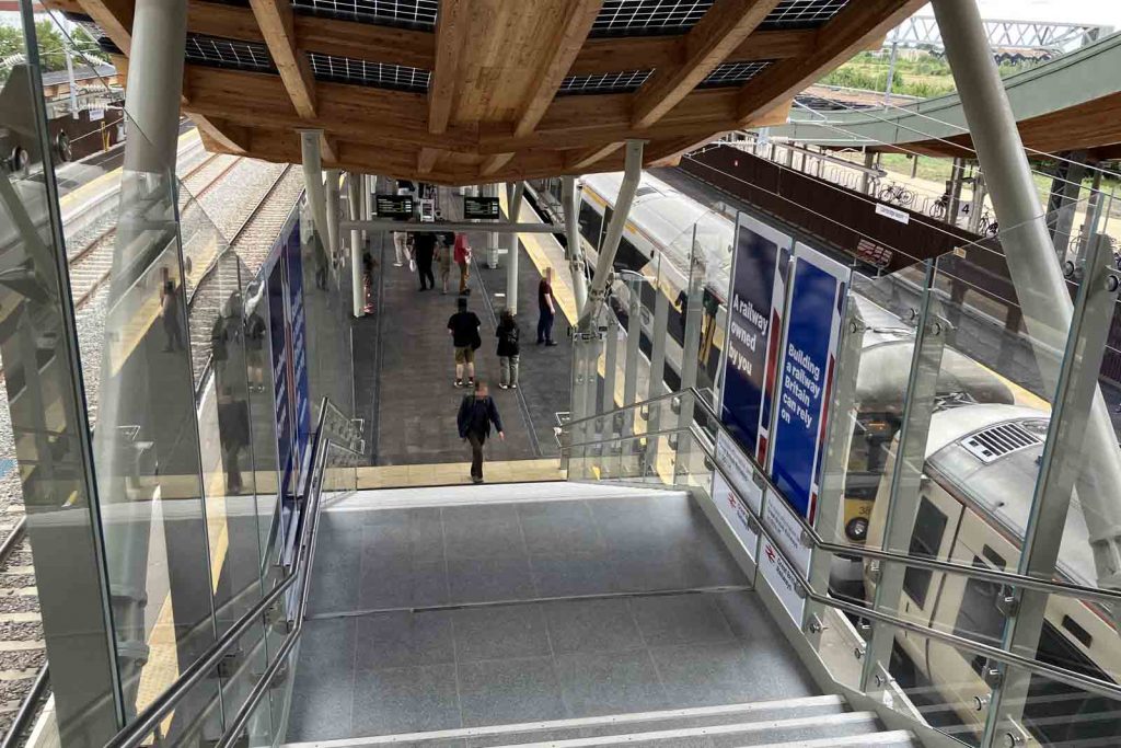 View down the steps to the island platform at Cambridge North station. The side walls are glass. On the intermediate landing, the wall panels have GBR posters with slogans "A railway owned by you" and "Building a railway Britain can rely on".