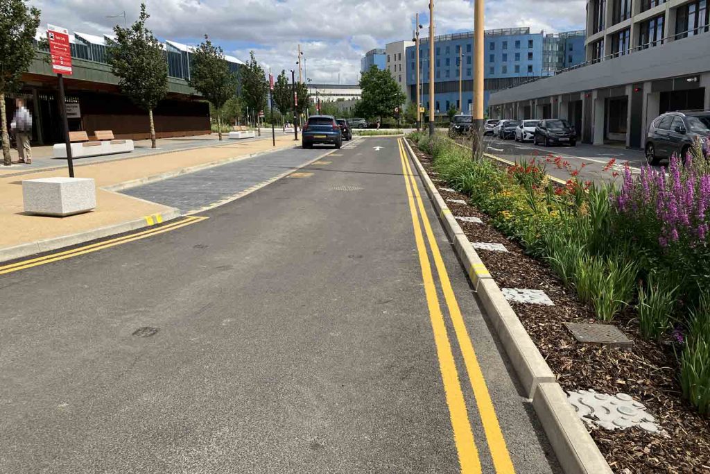 Photo looking along the approach road to Cambridge South station. Gaps in the kerb allow water to drain into the central reservation, which is planted with pretty flowers and has been mulched. Photo also shows the taxi rank, and large institutional buildings around the station.