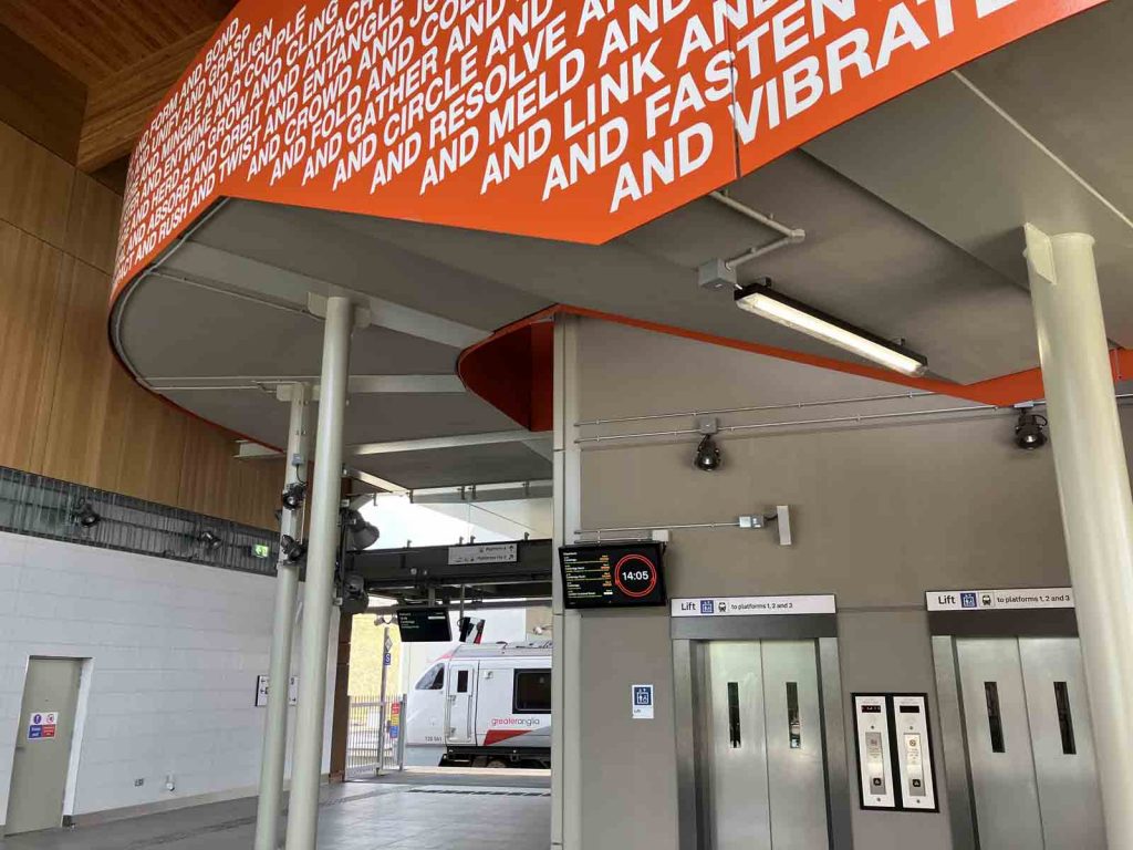 Photo inside the west side building at Cambridge South station, at ground level. We see the two lift doors, and the passageway to platform 4. A train is just seen at platform 3. We also see part of the outer parapet of the staircase as it curls around the lift shafts. On the bright orange parapet, there is text. The lines nearest to the camera, at the foot of the parapet, each begin as follows: "and circle... "and resolve... "and meld... "and link... "and fasten... "and vibrate..."