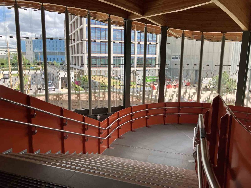 Photo looking down one of the staircases on the main footbridge at Cambridge South station. The parapets are steel panels, painted bright orange. The tall glass walls of the building structure allow us to see the institutional buildings nearby.